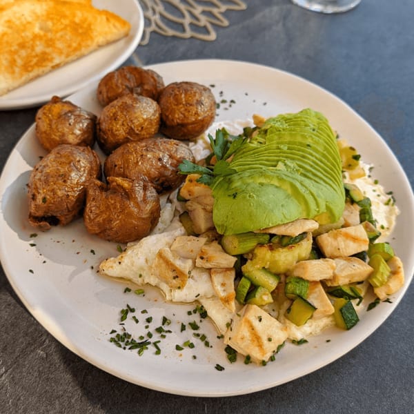 A plate of roasted potatoes, sliced avocado, chicken, zucchini, and herbs, with toast in the background.