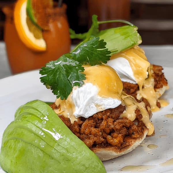 Chorizo eggs Benedict with hollandaise sauce, cilantro, and avocado, with a drink in the background.