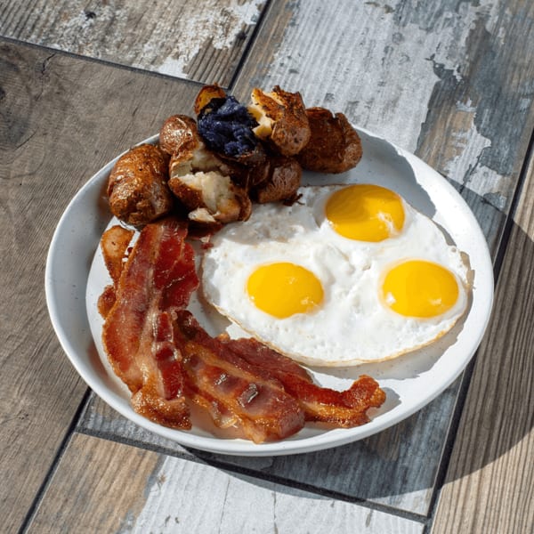 A breakfast plate with three fried eggs, crispy bacon, and roasted potatoes, one with purple flesh.